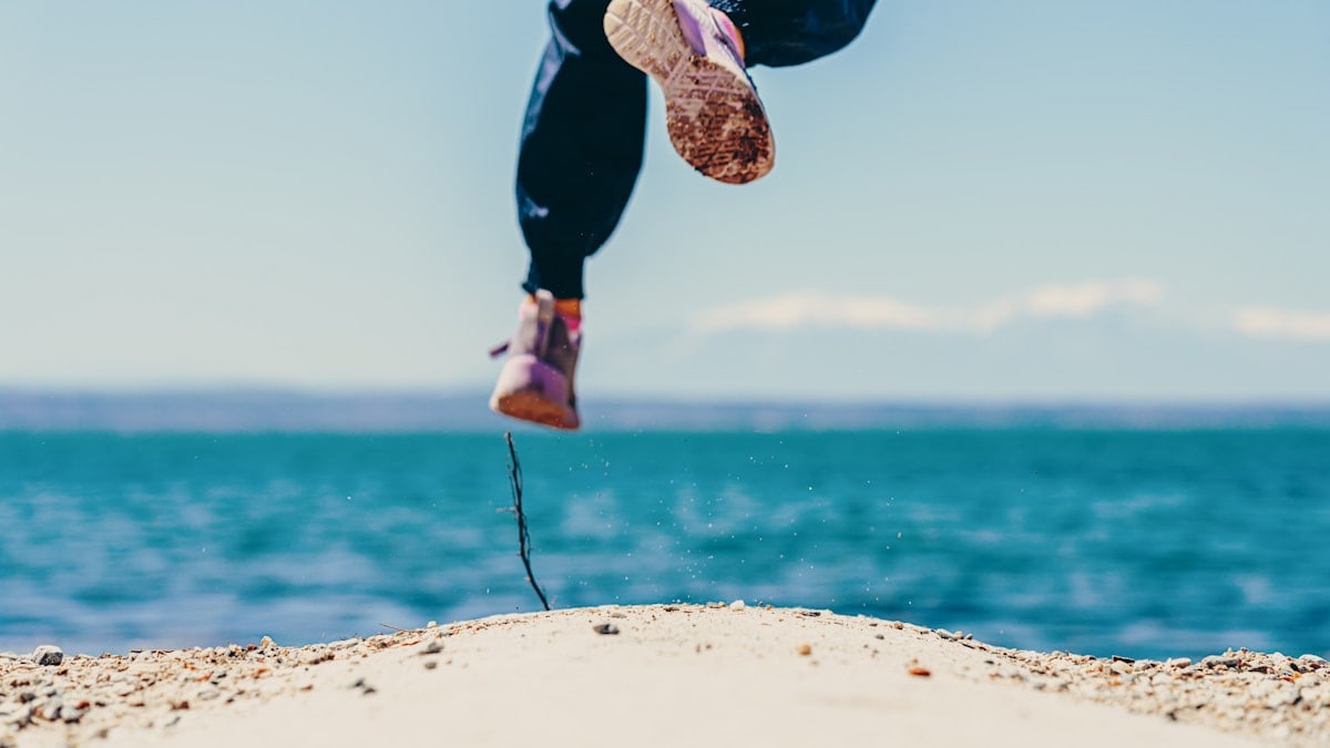 person in black pants and brown shoes jumping on brown rock near body of water during